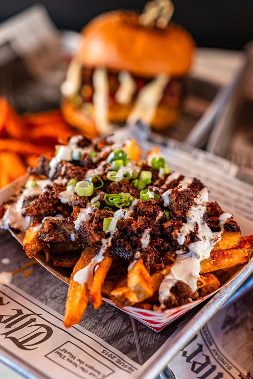 Loaded fries with meat, white sauce, and green onions, with a burger in the background.