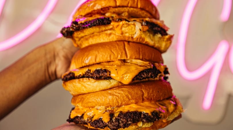 A hand holds a towering stack of four cheeseburgers with melted cheese and buns, against a pink neon sign.