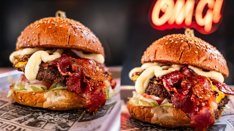 Two juicy burgers with bacon, cheese, lettuce, and mayonnaise, with a neon sign in the background.