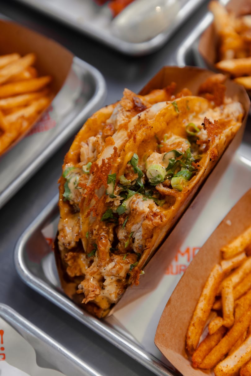 A crispy chicken taco with sauce and green onions, served with fries on a metal tray.