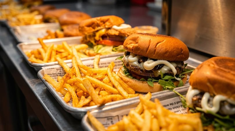 Multiple trays of gourmet burgers with melted cheese, arugula, and fries on a counter.
