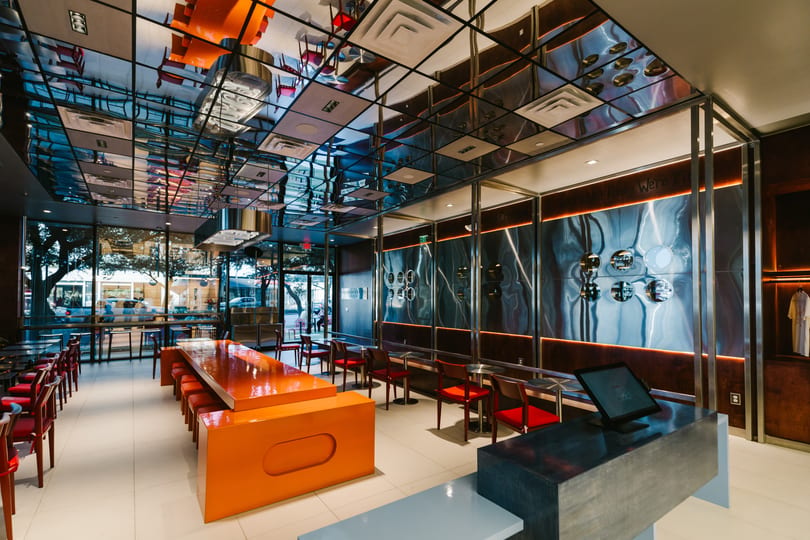 Modern restaurant interior with orange communal table, red chairs, and a reflective ceiling.