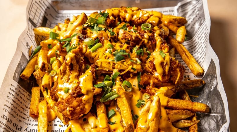 Loaded fries with cheese sauce, shredded cheese, chicken, and green onions in a newspaper-lined basket.