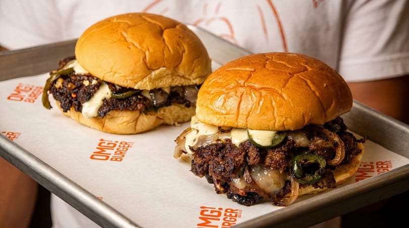 Two delicious OMG! Burgers with melted cheese, grilled onions, and jalapeños on a metal tray.
